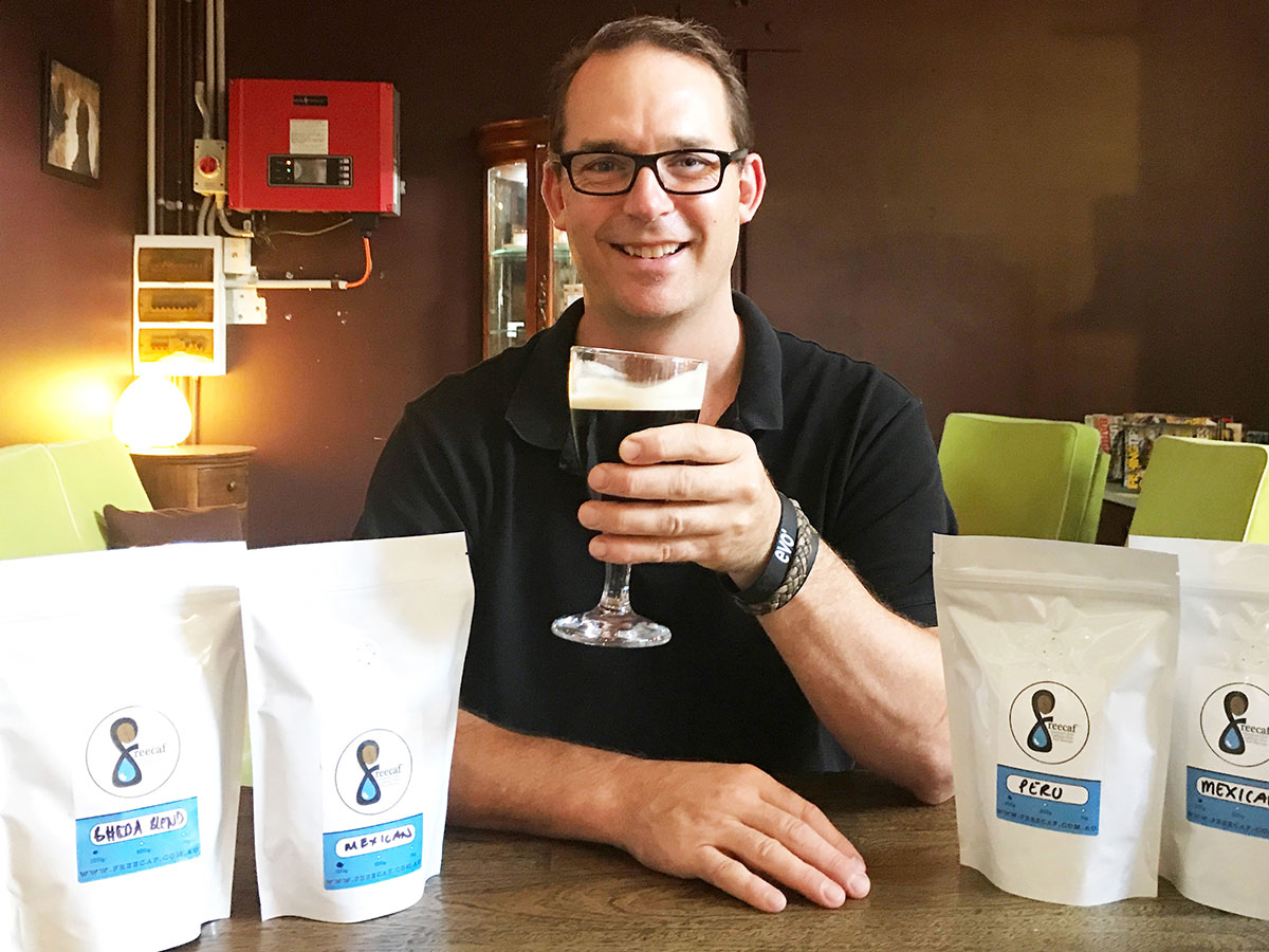 Daniel Salter holding a glass of nitro coffee in his hand while sitting at a table with bags of Freecaf Coffee beside him