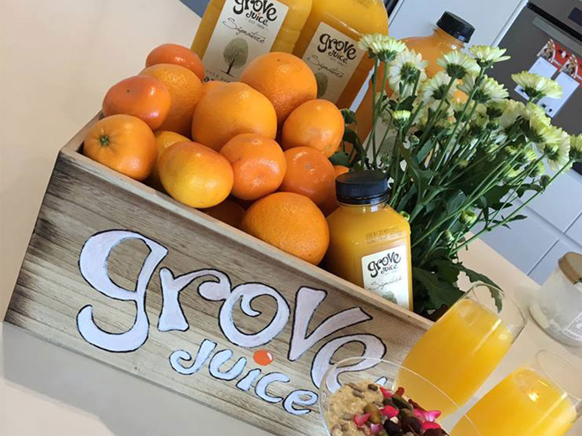 Box of oranges and pale yellow flowers with Grove Juice written on the side and two glasses of orange juice in the right hand corner.