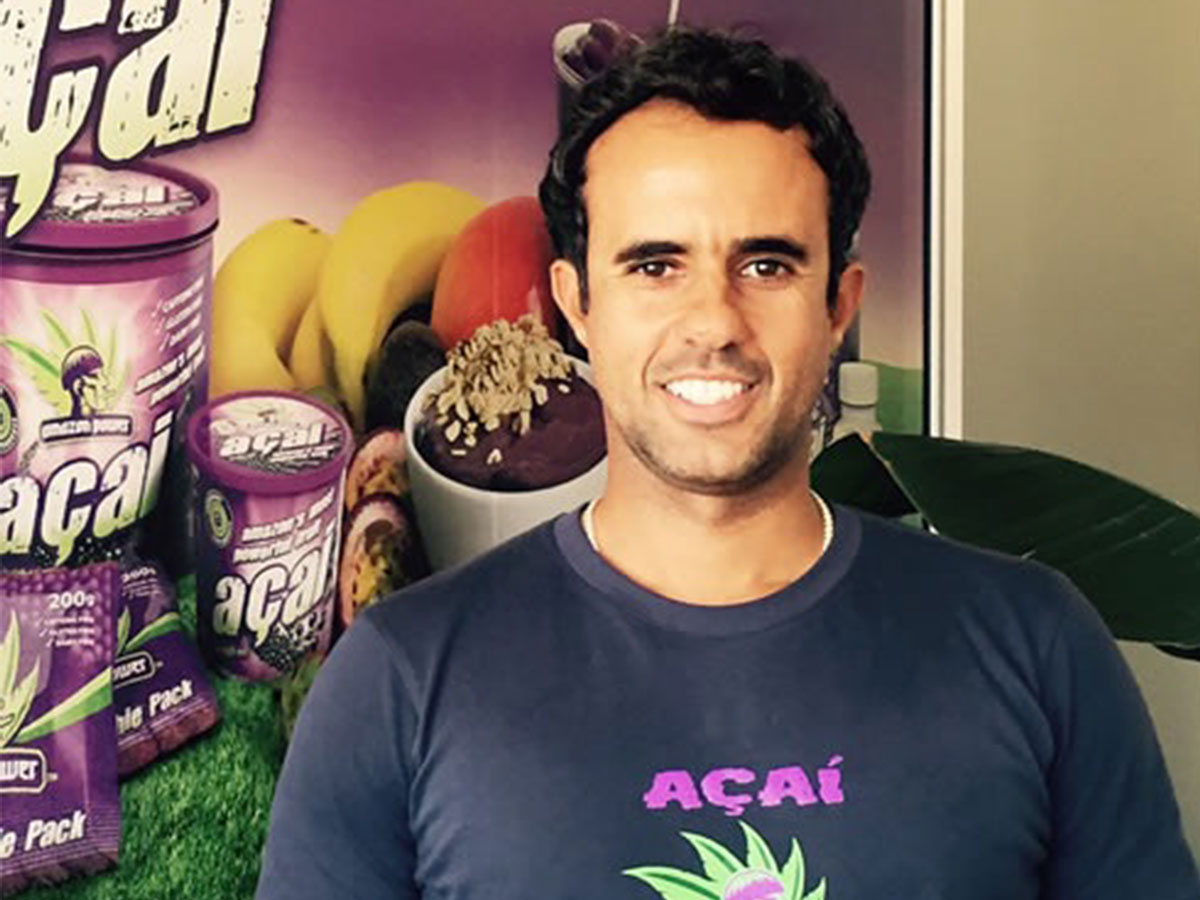Americo Tognetti, the original Acai man, dressed in a navy T shirt with Acai written on the font and a poster showing his Acai products in the background.