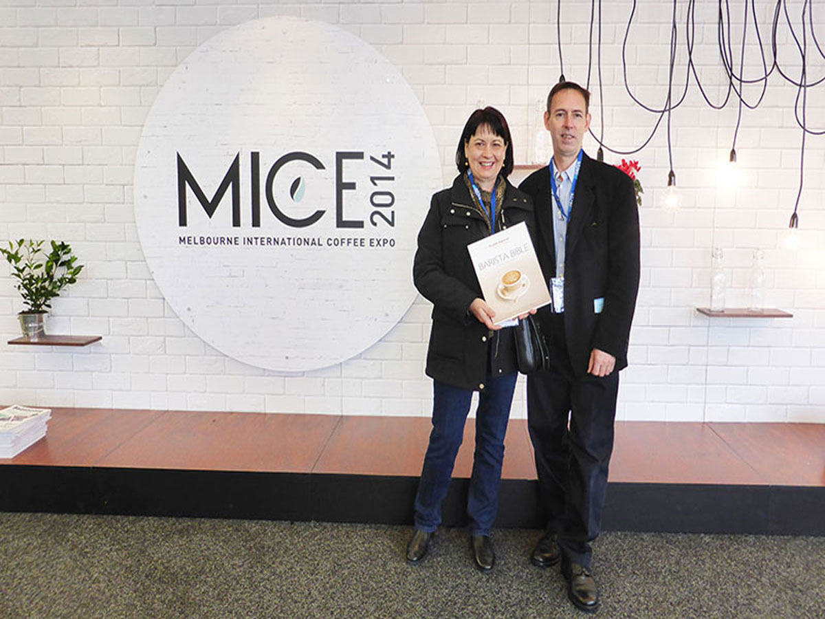 Paul Holiday & Christine Cottrell with Barista Bible near sign at entry of MICE 2014