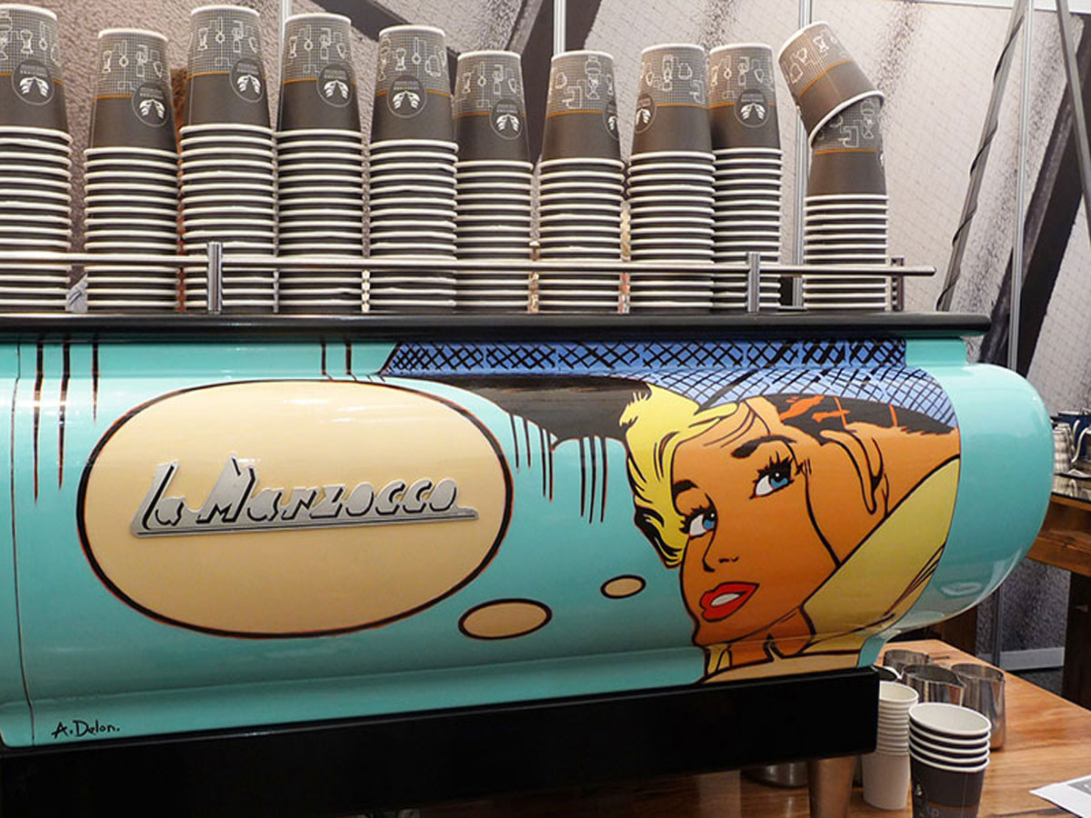 La Marzocco espresso machine with grey paper cups on top and comic characters painted on the side at MICE 2012