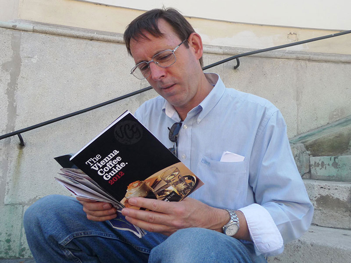 Paul Holiday dressed in a blue shirt and jeans reading the book The Vienna Coffee Guide 2012 published by Allegra Strategies - sitting on steps outside a cafe in Vienna