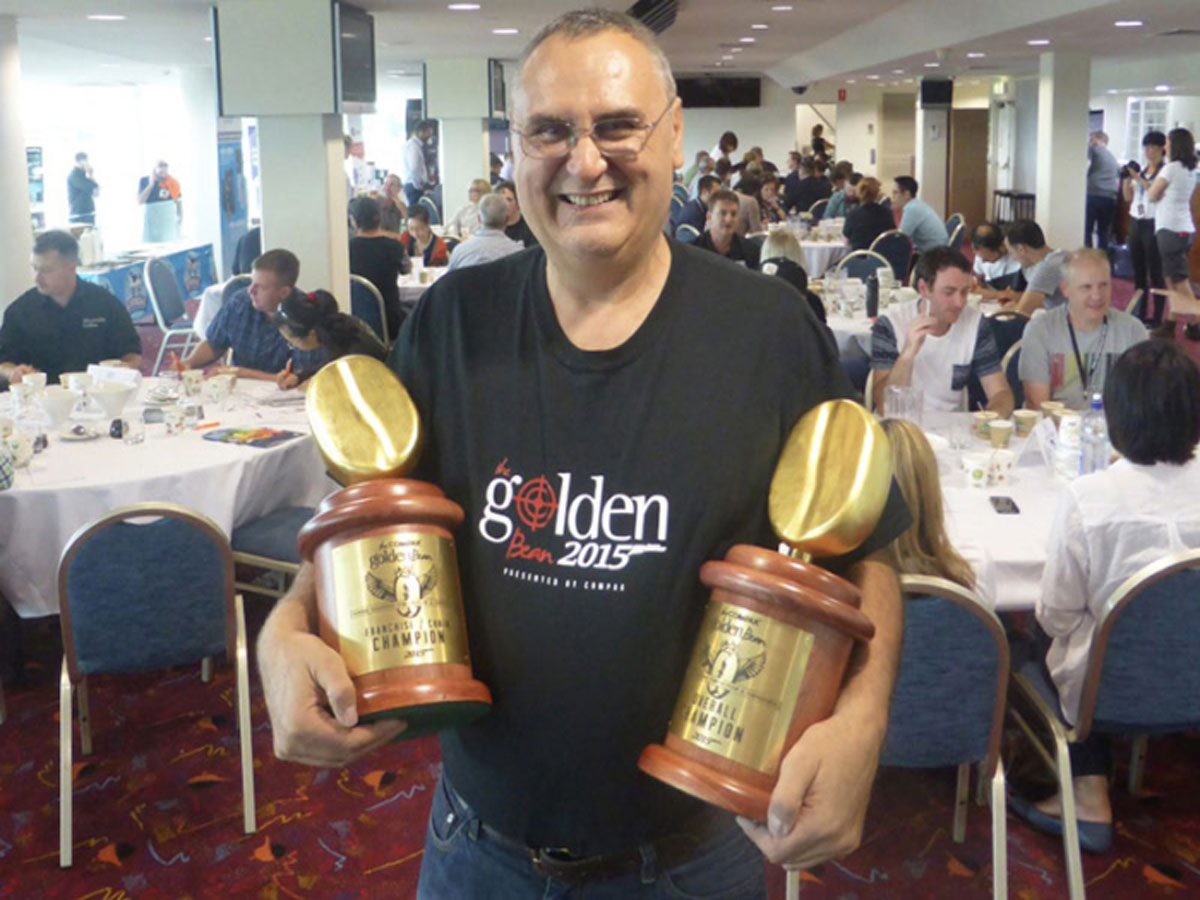 Reg Barber holding two trophies at 2015 Golden Bean in Newcastle - with coffee judging in the background