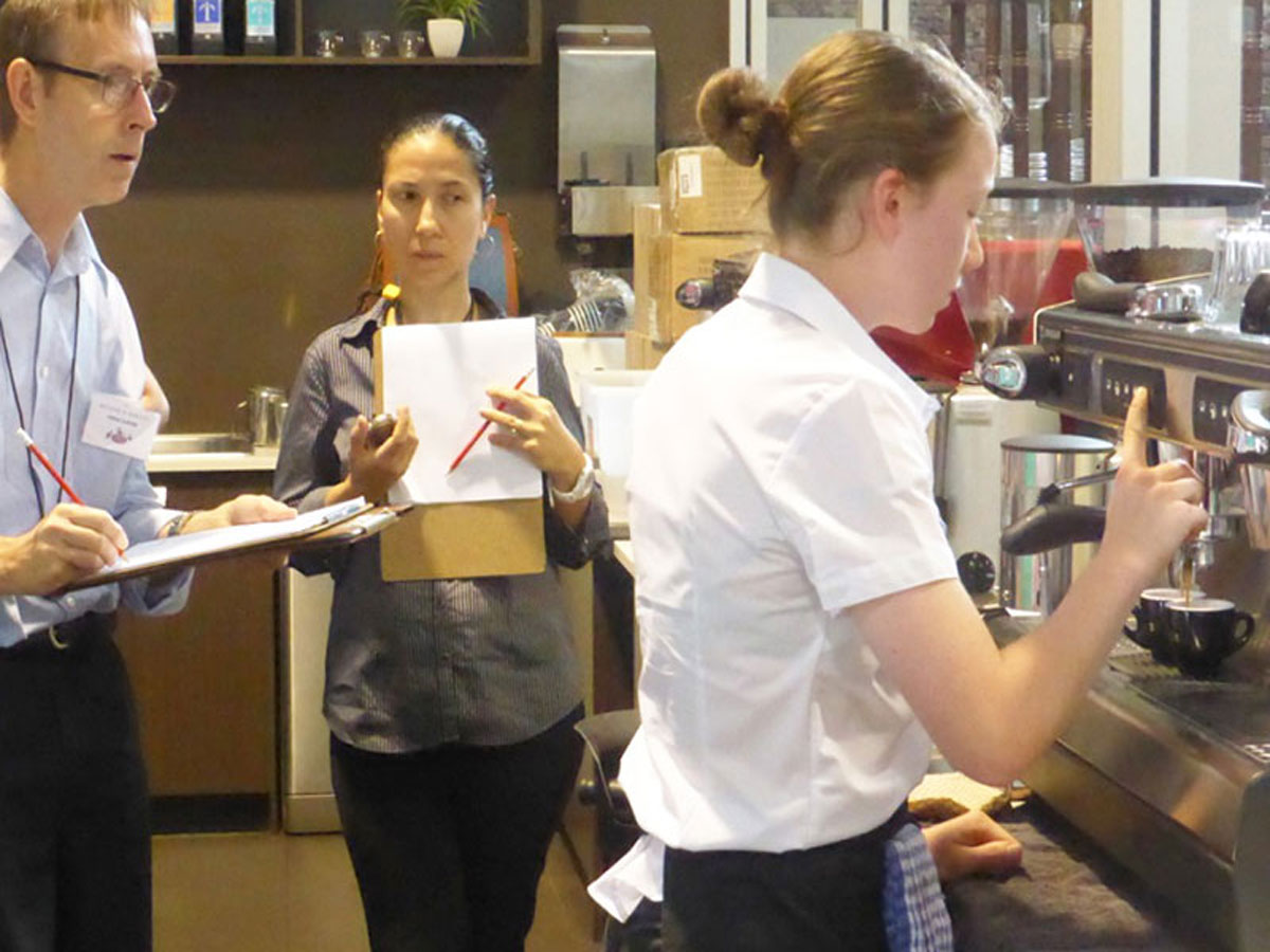 Paul Holiday and Carla De Jesus holding score sheets and pencils judging a student making espresso at the 2016 Become A Barista competition held at Grinders Coffee, Teneriffe