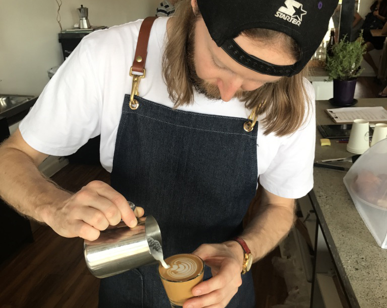 Barista from Valdmeer Cafe with long hair and a black apron and cap on backwards pouring milk into a glass making latte art