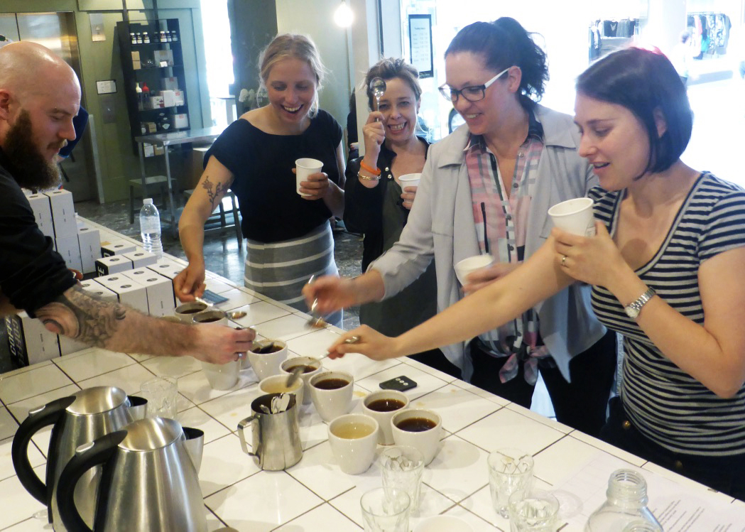 Hannah Hutonen, Lisa Feely, Anne Cooper and Georgia Major cupping coffee standing at a counter discussing the coffee with a barista on the other side at Singe Origin in Melbourne.